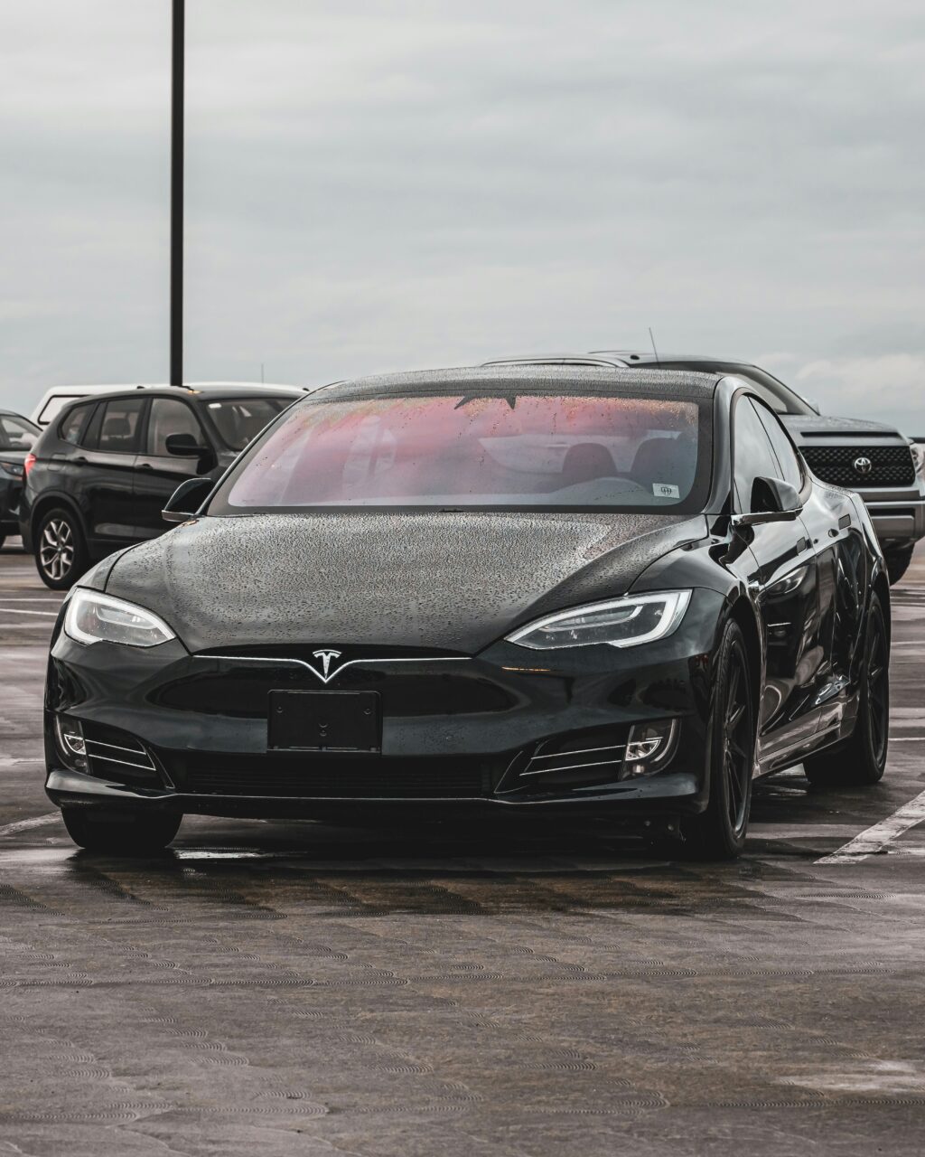Front view of a black Tesla Model S covered in rain droplets, parked in a public lot on a cloudy day.