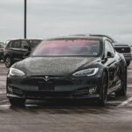 Front view of a black Tesla Model S covered in rain droplets, parked in a public lot on a cloudy day.