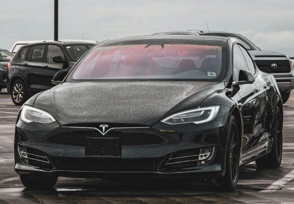 Front view of a black Tesla Model S covered in rain droplets, parked in a public lot on a cloudy day.