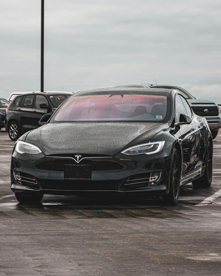 Front view of a black Tesla Model S covered in rain droplets, parked in a public lot on a cloudy day.